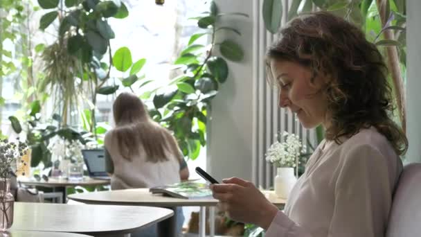 Szczęśliwa młoda kobieta teksty na telefon siedzi w stylowej kawiarni - Materiał filmowy, wideo