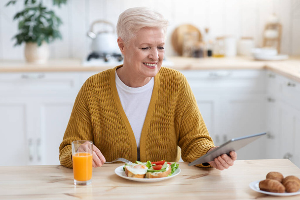 Mujer mayor positiva almorzando y usando tableta digital - Foto, Imagen
