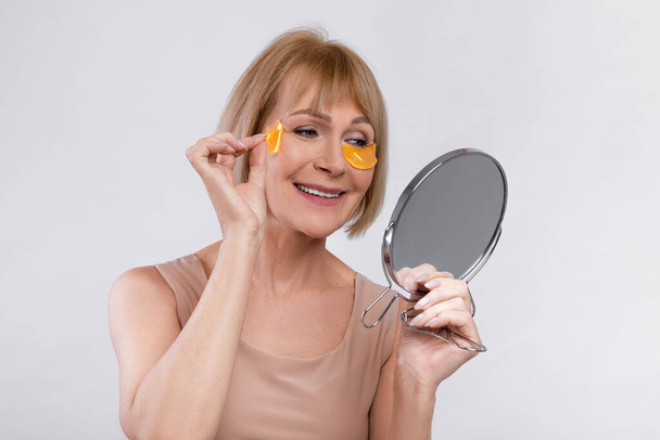 Glimlachende volwassen dame met behulp van gouden hydrogel oogvlekken in de buurt spiegel over lichte studio backgroud. Schoonheid en huidverzorging - Foto, afbeelding