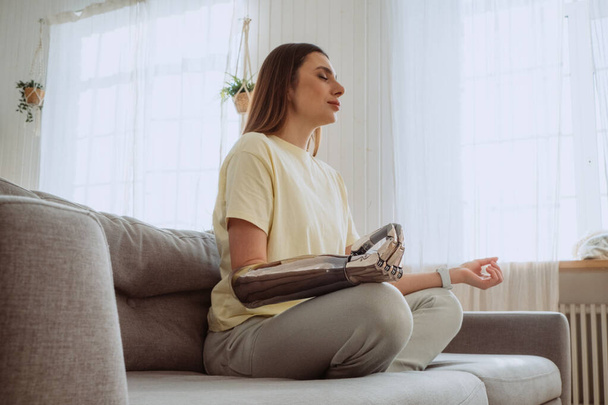 Blonde Frau mit stylischem Prothesenarm meditiert auf Sofa - Foto, Bild