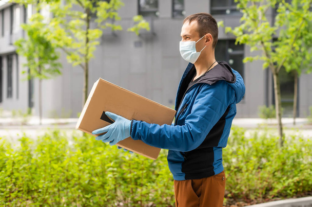 Курьер почтовой доставки, одетый в защитную маску перед грузовым фургоном, доставляет коробку с коронавирусной болезнью или COVID-19 - Фото, изображение