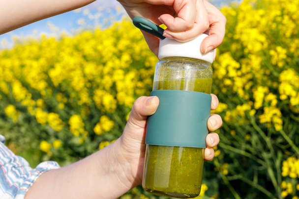  女は用意された緑色のスムージーのガラス瓶を開ける。健康的,混合果物や野菜の飲み物,背景,黄色の花を咲かせます,自然と調和した生活の概念,適切な栄養  - 写真・画像
