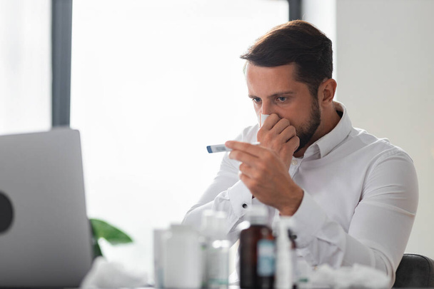 Young male businessman blows his nose and measures the temperature with a thermometer while sitting in the office, Influenza virus, cold, feeling unwell concept - Fotó, kép