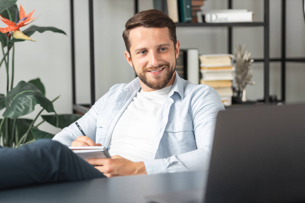 職場や家庭のオフィスでオンライン会議のためのラップトップを使用してカジュアルな服で陽気な成功した若い男は、机の上に足を保持しています - 写真・画像