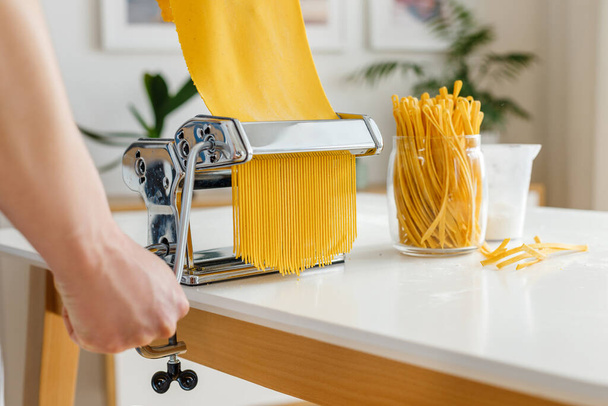 麺カッターでスパゲティを作るエプロンの男。自宅でのクローズアップパスタ料理。鮮やかな黄色の生地から食品を準備. - 写真・画像