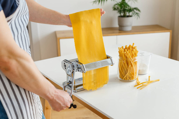麺カッターでスパゲティを作るエプロンの男。自宅でのクローズアップパスタ料理。鮮やかな黄色の生地から食品を準備. - 写真・画像