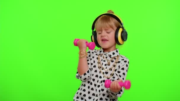 Dziewczynka słucha muzyki przez słuchawki, ćwiczy pompowanie mięśni ramion podnosząc różowe hantle - Materiał filmowy, wideo