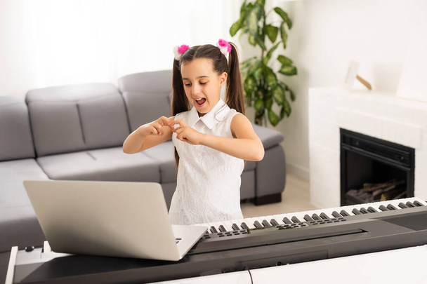 家庭学校の小さな子供の女の子は、学校の先生によってインターネット音楽オンラインクラスに接続するコンピュータからピアノを学ぶ。新しい普通の生活と教育、家庭の概念での学生の研究. - 写真・画像