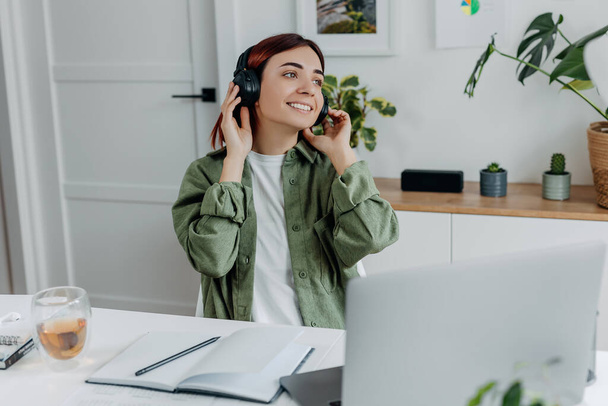 Woman listening music with wireless headphones. Young businesswoman relaxing at comfortable workplace. Concept of resting during online business at home office,telework or studing. Happy girl singing. - Foto, Bild