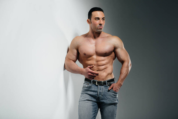 A young athlete bodybuilder poses in the studio topless in jeans near the wall. Sport. - Foto, afbeelding