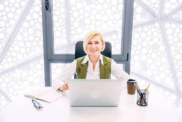 Atractiva rubia jefa gerente se sienta en su oficina en el lugar de trabajo, utilizando el ordenador portátil, sonrisas y bebidas café - Foto, Imagen