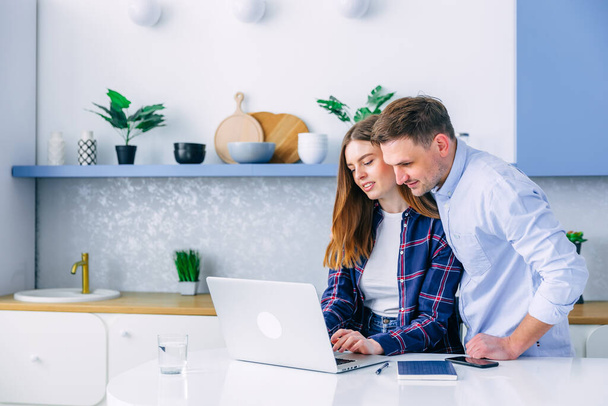 Young caucasian couple of woman and man sitting at home kitchen looking to computer laptop screen smiling laughing pointing to screen excited cheerful happy. Couple buying products or services online - Φωτογραφία, εικόνα