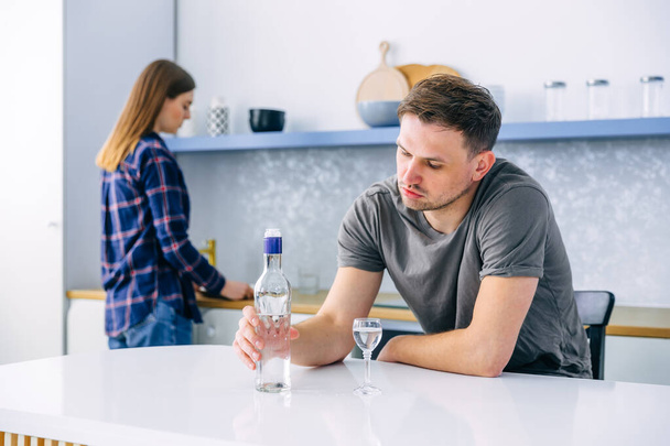 Σοβαρά δυστυχισμένος άνθρωπος κάθεται στο τραπέζι και ρίχνει βότκα σε ένα ποτήρι για τον εθισμό στο αλκοόλ. Η απελπισμένη σύζυγος στέκεται με την πλάτη της. αλκοολισμός στην οικογένεια - Φωτογραφία, εικόνα