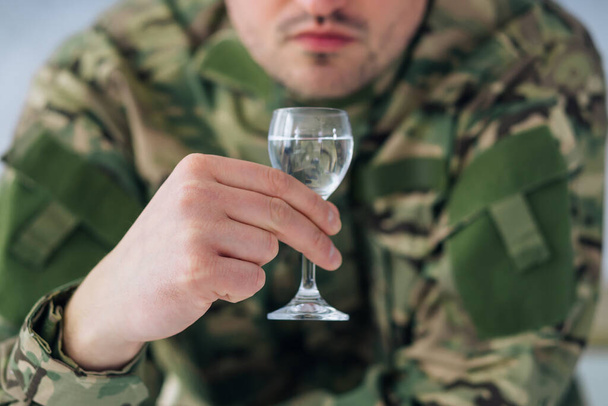 manos de jóvenes soldados en uniformes de camuflaje sentados en casa con una botella de vodka bebida alcohólica sosteniendo un vaso. Depresión después del servicio militar, alcoholismo y adicción - Foto, imagen