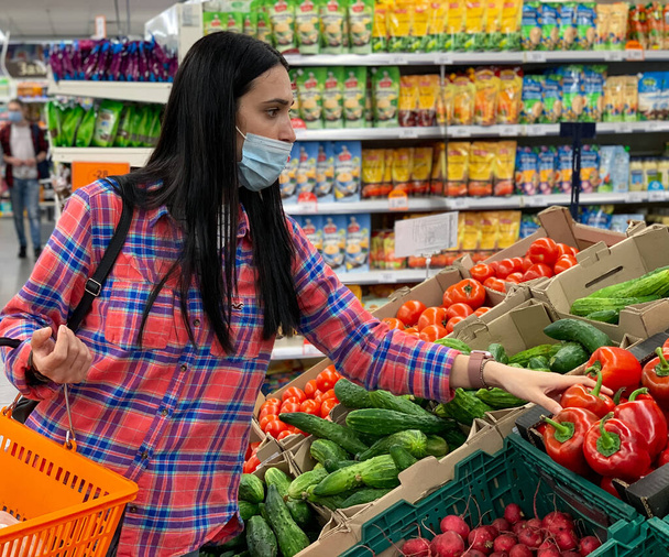 スーパーで野菜を選ぶ買い物かご付きの医療用マスクの女性。コロナウイルスの流行からの保護,新鮮な果物と免疫力の増加.ウイルスと戦うための健康食品. - 写真・画像