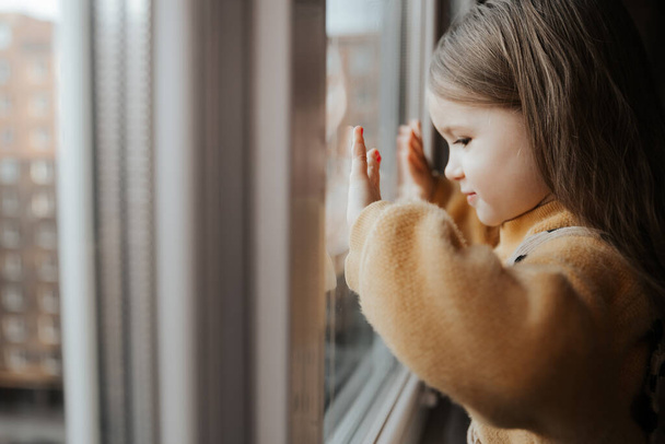 Ein kleines Mädchen auf der Fensterbank blickt aus dem Fenster der Wohnung in einem Hochhaus - Foto, Bild