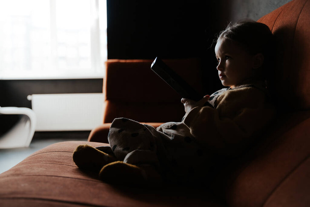 Photo of little girl using remote control and holding copyspace while watching tv on couch at home - Foto, Bild