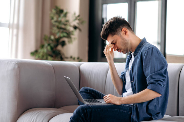 Upset tired young caucasian bearded man sitting on sofa in living room, in casual clothes, using laptop, student in stress, overworked with study and homework, rubs eyes, needs rest - Фото, зображення