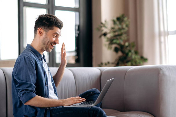 Счастливый дружелюбный молодой кавказский бородач, сидящий на диване, одетый в стильную джинсовую одежду, пользующийся ноутбуком, общающийся с друзьями или семьей через видеоконференцию, машущий рукой, улыбающийся - Фото, изображение