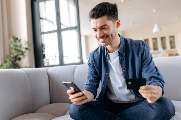 Homem caucasiano feliz e bem sucedido sentado no sofá na sala de estar, vestido com roupas casuais, usando smartphone e cartão de crédito para compras on-line, digitando dados para pagamento, sorrindo - Foto, Imagem