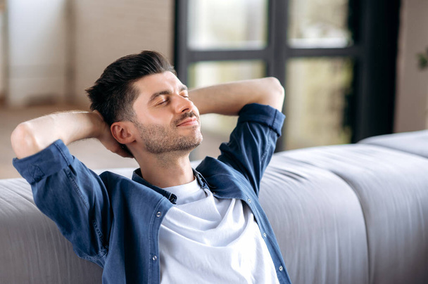Close-up calm happy handsome caucasian bearded man in casual clothes relaxing on sofa at home, resting in living room, dreaming of vacation, put his hands behind his head, close his eyes, smiling - Φωτογραφία, εικόνα