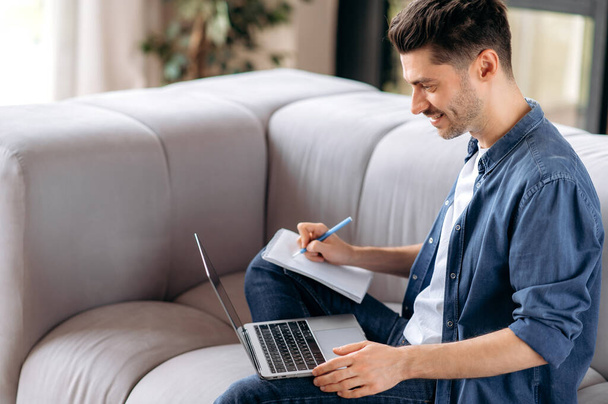 Smart confident young caucasian man sitting on the couch, in casual clothes, student doing online distance learning, group brainstorming, taking notes, looking at the screen, smiling - Fotó, kép