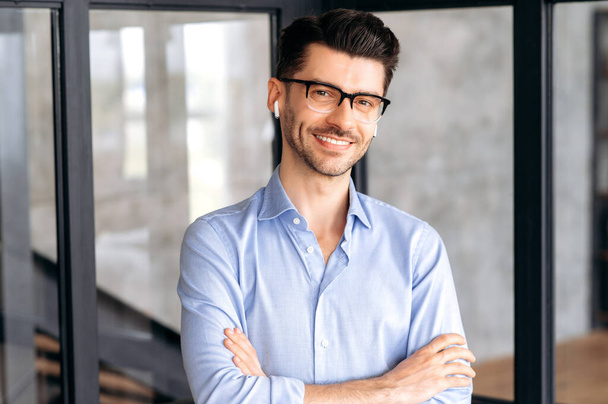 Portrait of joyful attractive confident young adult business man or manager wearing glasses and headphones, wearing formal shirt, standing in office, looking at camera, arms crossed, smiling friendly - Foto, Imagen