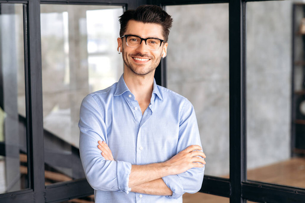 Portrait successful confident smart young adult business man or manager wearing glasses and headphones, wearing stylish clothes, standing in office, looking at camera, arms crossed, friendly smiling - Photo, Image