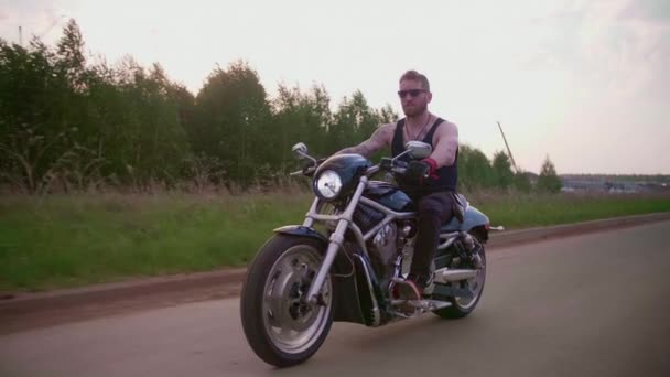 Elegante motociclista con tatuajes monta una motocicleta en una carretera de campo al atardecer - Metraje, vídeo
