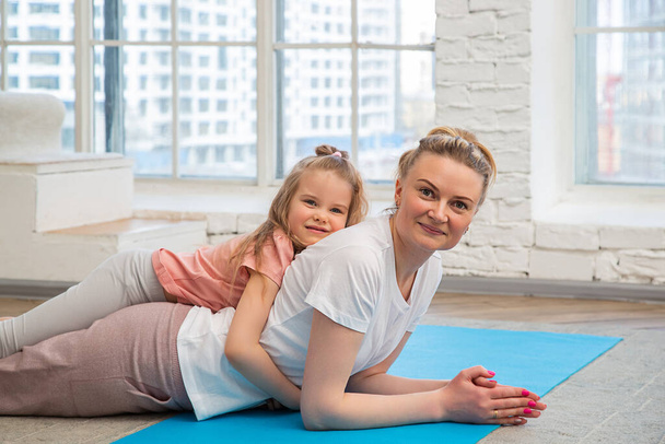 Portrait of mother with her preschool-aged caucasian daughter in sportswear lying on a yoga mat in front of living room window, hugging and looking at the camera. Spending time with children at home. - Fotó, kép