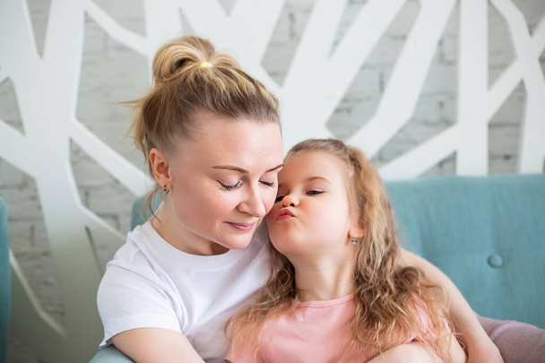 Little caucasian girl sitting together with mother on sitting on the sofa in living room, hugging and kissing each other. Spending time with children at home. - Фото, зображення