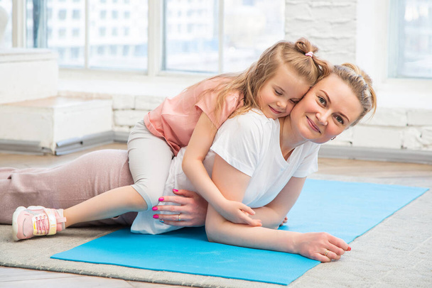 Portrait of mother with her preschool-aged caucasian daughter in sportswear lying on a yoga mat in front of living room window, hugging and looking at the camera. Spending time with children at home. - Foto, imagen