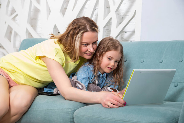 Portrait of mother with her preschool-aged caucasian daughter lying on the couch with a laptop and look at the monitor with a smile. Online learning concept, spending time with children at home. - Foto, Bild