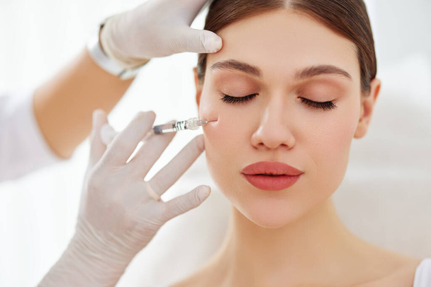 Mujer joven que recibe la inyección en el pómulo de cosmetólogo profesional en máscara y guantes en el procedimiento de belleza en la clínica moderna - Foto, Imagen