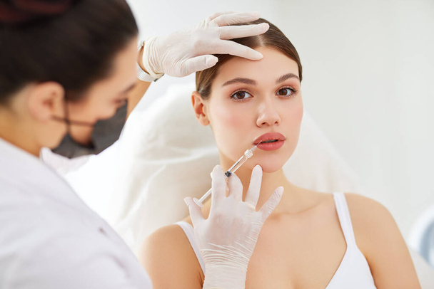 Genç bayan hasta, modern klinikte maske ve eldiven takan profesyonel kozmetik uzmanının dudaklarına enjekte ediliyor. - Fotoğraf, Görsel