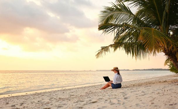 Pracuje na dálku na pobřeží. Mladá úspěšná žena žena na volné noze v slamáku pracuje na notebooku, zatímco sedí na tropické pláži při západu slunce, v celé délce. Koncept práce na dálku - Fotografie, Obrázek