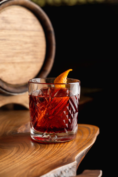 American old fashioned cocktail garnished with an orange zest, wooden barrel on a background, unusual creative wooden table - Foto, Imagem