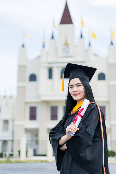 Eine junge schöne asiatische Universitätsabsolventin im Abschlusskleid und mit einem Diplom steht vor dem Universitätsgebäude, nachdem sie am College-Start teilgenommen hat - Foto, Bild