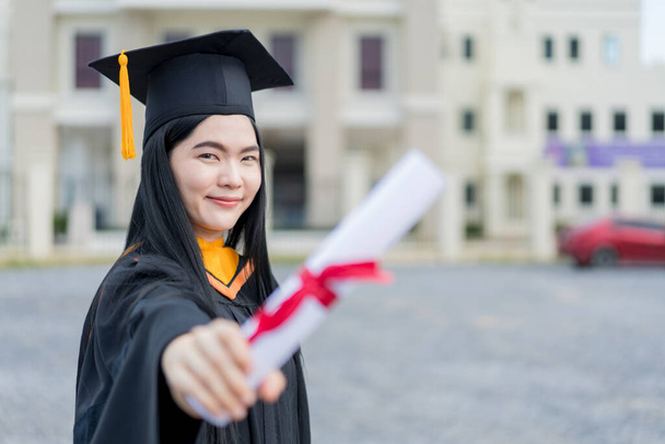 A young beautiful Asian woman university graduate in graduation gown and mortarboard holds a degree certificate stands in front of the university building after participating in college commencement - Valokuva, kuva