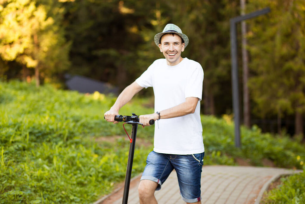 Portret van een jonge knappe man in wit shirt, zonnebril en hoed klaar om zijn elektrische scooter te rijden op straat in de zonnige zomer. Ecologisch transportconcept. Elektrisch vervoer. - Foto, afbeelding