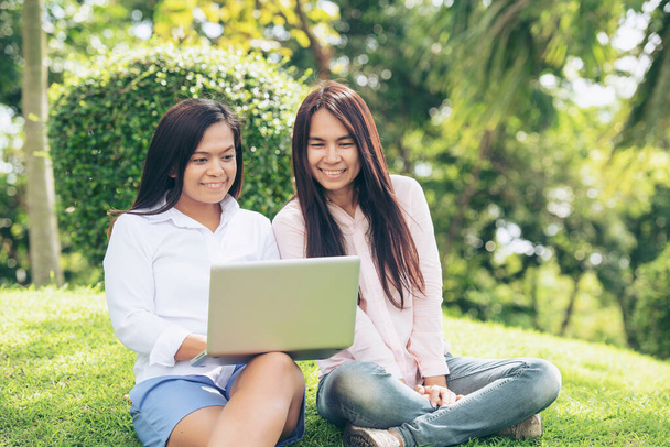 Asian two women talking using laptop sitting on green meadow at park. Friendship girls chat and talk look at laptop planning do small business green garden. Young beautiful women laugh talk together. - Foto, Imagen