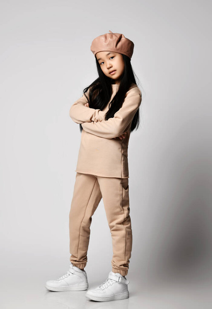 Cool asian brunette kid girl in leather hat beret, cotton pants, hoodie, jersey and sneakers stands with arms crossed - Foto, Bild
