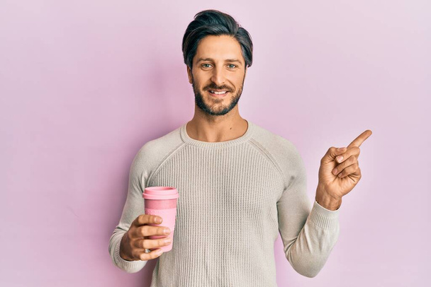 Young hispanic man drinking a take away cup of coffee smiling happy pointing with hand and finger to the side  - Фото, зображення