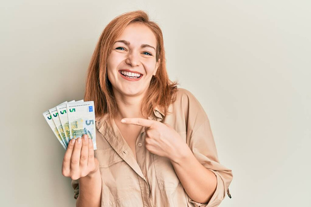 Young caucasian woman holding bunch of 5 euro banknotes smiling happy pointing with hand and finger  - 写真・画像