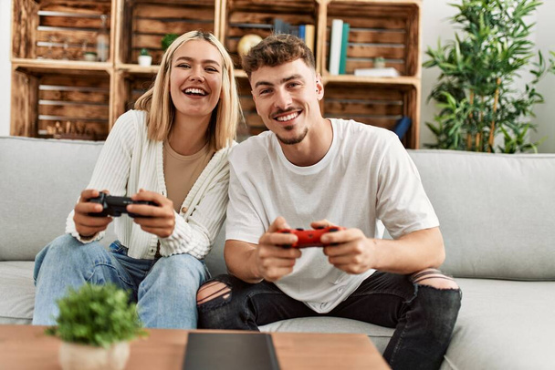 Νεαρό καυκάσιο ζευγάρι χαμογελά χαρούμενος παίζοντας βιντεοπαιχνίδι στο σπίτι. - Φωτογραφία, εικόνα