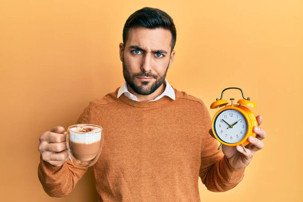 Junger hispanischer Mann, der Kaffee und Wecker in der Hand hält, ist skeptisch und nervös und runzelt ob des Problems die Stirn. negative person.  - Foto, Bild