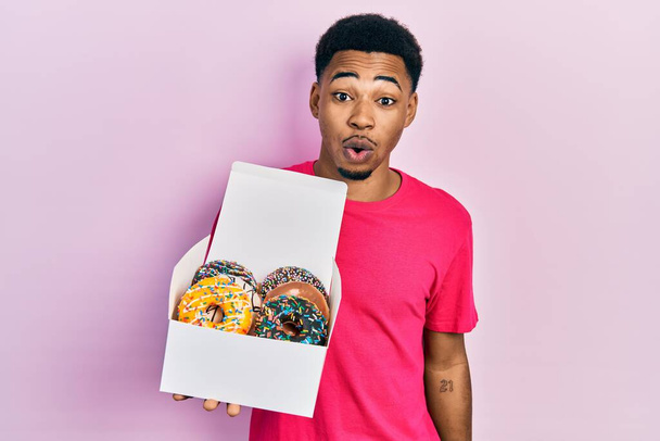 Mladý africký Američan drží krabici s chutnými barevnými koblihami vystrašené a ohromen s otevřenými ústy pro překvapení, nevěřící tvář  - Fotografie, Obrázek