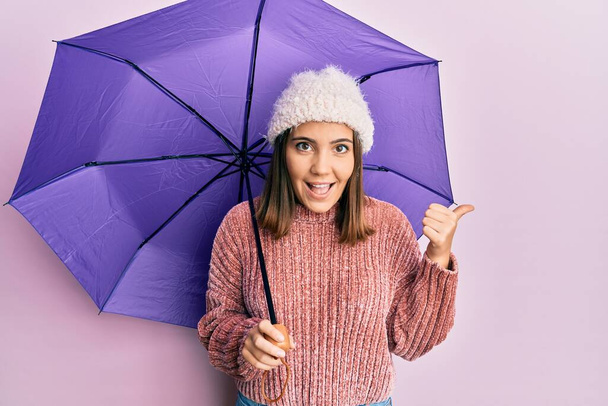 Young beautiful woman holding purple umbrella pointing thumb up to the side smiling happy with open mouth  - Foto, imagen