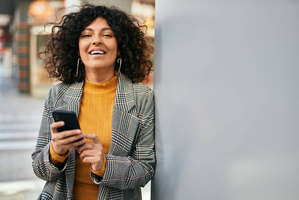 Giovane donna d'affari ispanica sorridente felice utilizzando smartphone in città. - Foto, immagini