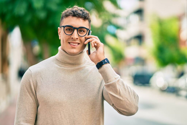 Νεαρός Ισπανός που φοράει γυαλιά και μιλάει σε smartphone στην πόλη. - Φωτογραφία, εικόνα